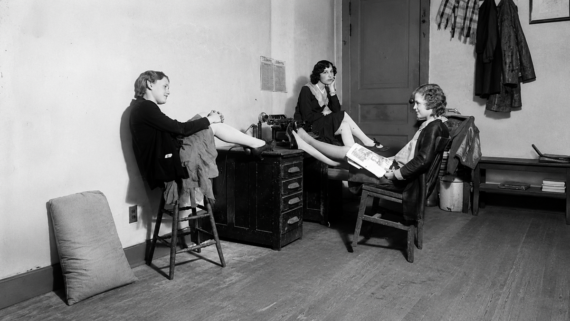 Three women sit in a sparsely furnished room, each with their legs up on a central desk. Two sit on stools and one on a chair, reading a magazine, while coats hang on the wall behind them.