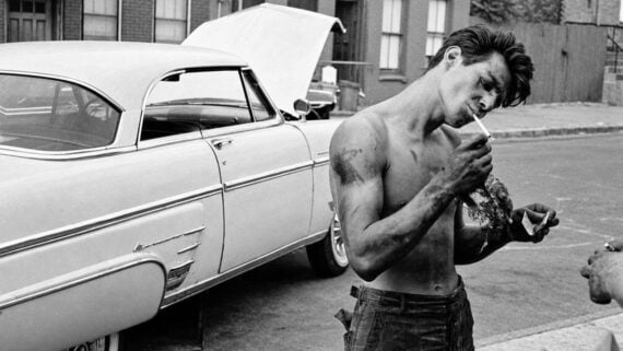 A shirtless, grease-stained man lights a cigarette while holding a rag next to a classic car with its hood up.