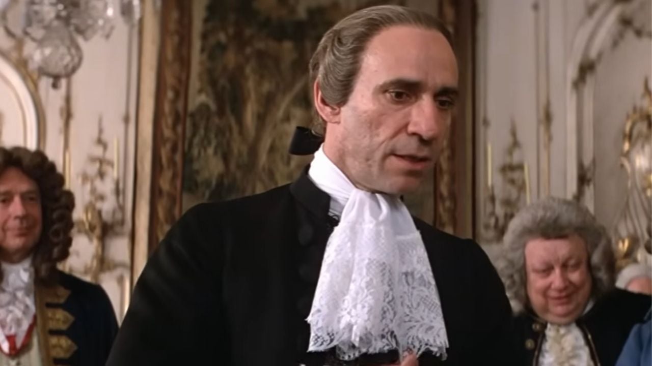A man in 18th-century attire with a white lace cravat stands indoors, with two other men in similar period clothing in the background. Ornate decor and a tapestry are visible behind them.