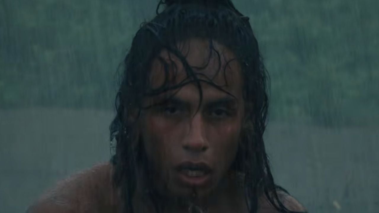 A person with wet hair stands in heavy rain, looking forward intensely. The background is blurred and green, suggesting an outdoor setting.