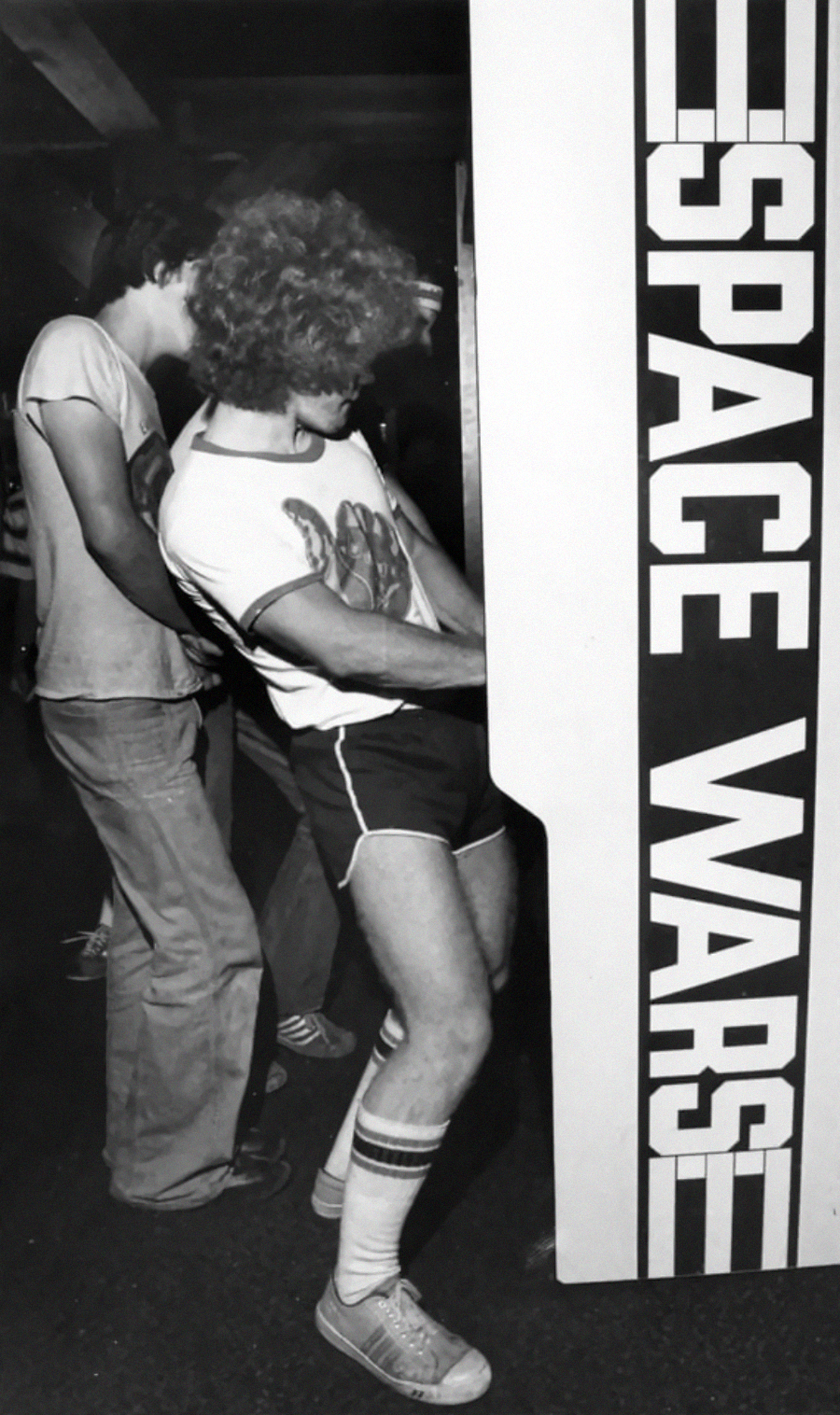 Two people play an arcade game called "Space Wars." The person in front wears a curly wig, a patterned t-shirt, shorts, tube socks, and sneakers, leaning enthusiastically toward the machine.