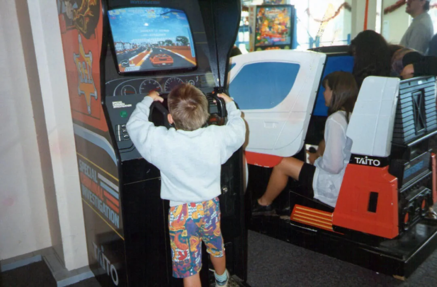 A young boy in colorful shorts plays a racing arcade game, holding a steering wheel. Nearby, other children are seated in vintage arcade machines. The scene is lively and nostalgic.