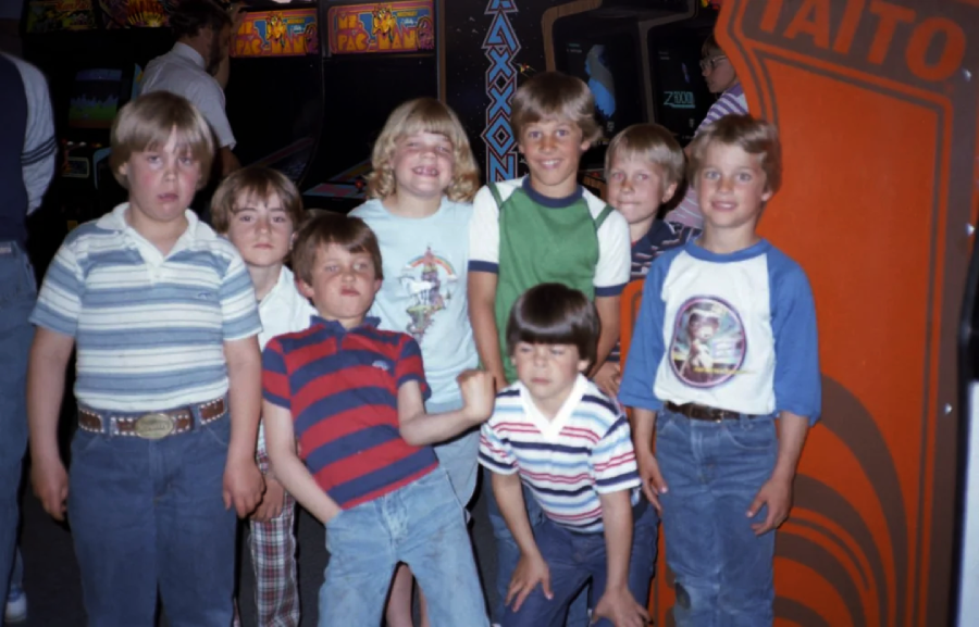 A group of eight smiling boys pose in front of vintage arcade machines, including "Pac-Man," "Zaxxon," and a Taito cabinet, in a lively arcade setting.