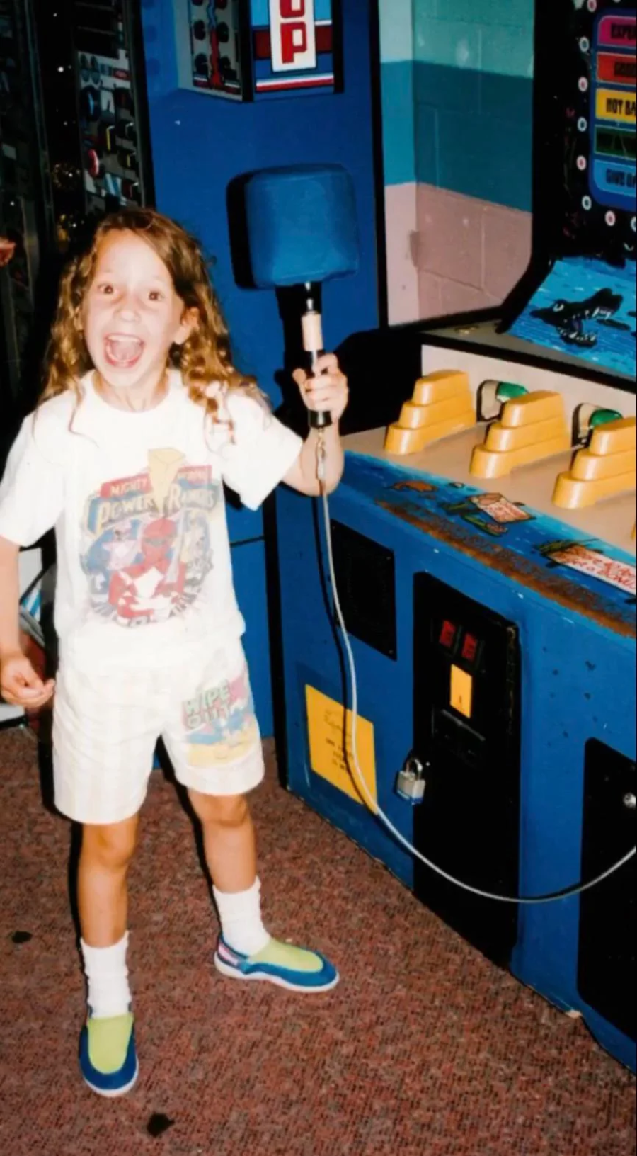 A smiling child with long curly hair holds a mallet at an arcade whack-a-mole game, wearing a Power Rangers T-shirt, shorts, and colorful sneakers. The arcade setting has blue and yellow machines.