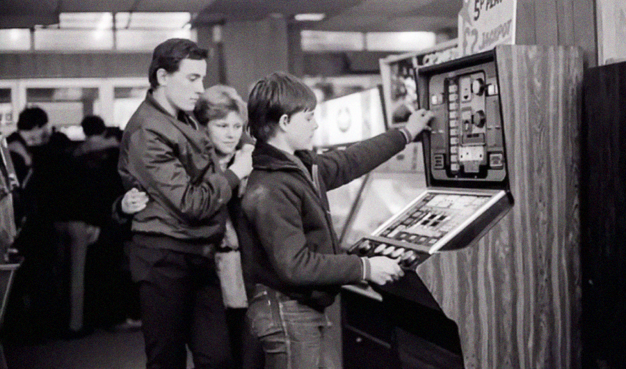 A black-and-white photo shows three teenagers in a retro arcade. One boy plays a vintage arcade game, while a couple stands behind him watching. The scene captures a nostalgic 1980s atmosphere.
