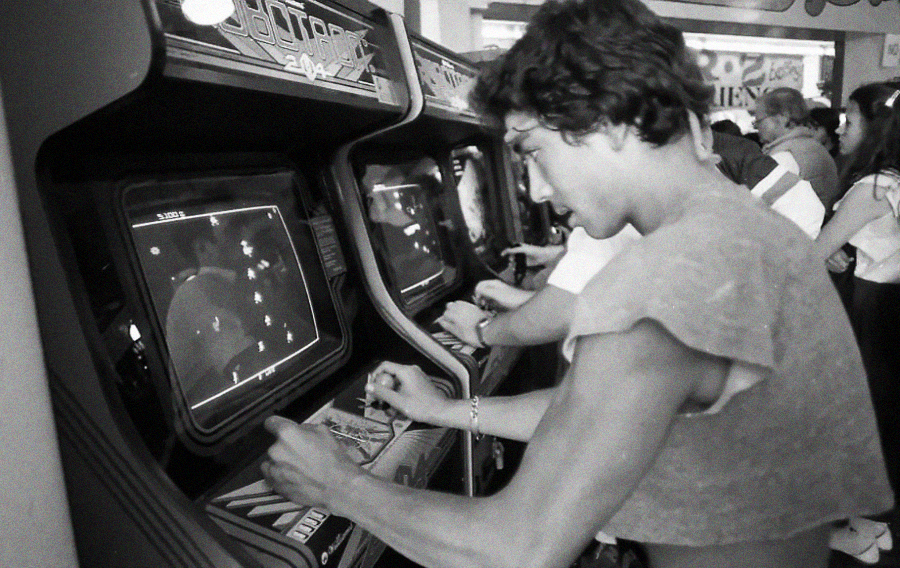 A young man plays a classic arcade game in a busy arcade, intensely focused on the screen, surrounded by other people also playing arcade machines. The photo is in black and white, evoking a retro feel.