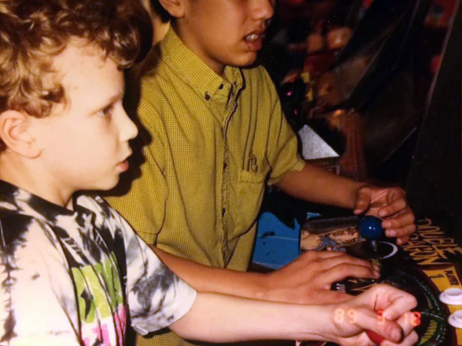 Two children intensely play an arcade video game, with one gripping a joystick and the other pressing buttons. The game’s controls and part of the screen are visible, and the photo has a nostalgic, vintage feel.