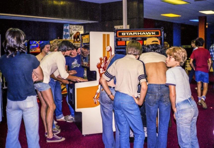 A group of boys and teens in 1970s or 1980s clothing crowd around arcade game machines in a retro game arcade with red carpet and colorful overhead lights.