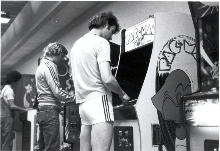 Black and white photo of three men playing arcade games. The man in the foreground stands at a Pac-Man machine, wearing a t-shirt and athletic shorts. The setting appears to be an arcade in the late 1970s or early 1980s.