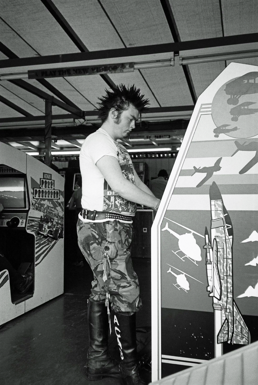 A person with a spiked mohawk, white T-shirt, camouflage pants, and tall boots plays an arcade game in a retro arcade setting. The scene has a vintage, 1980s atmosphere.