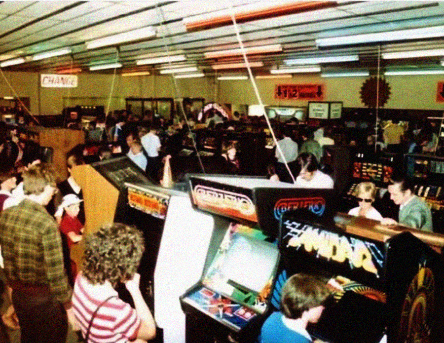 Crowded retro arcade filled with people playing and watching classic arcade games, including Centipede and Tempest, under bright fluorescent lights and a high ceiling.