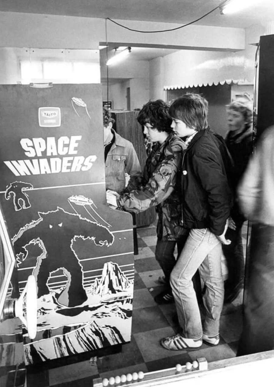 Several young people stand around and play a retro Space Invaders arcade game in a black-and-white photo, evoking a vintage atmosphere from the late 1970s or early 1980s.