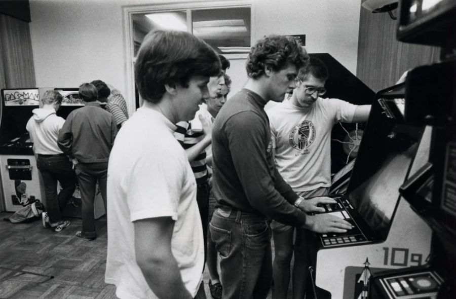 A group of young men in casual clothing play and watch arcade games in a retro-style arcade room, surrounded by various classic arcade machines.