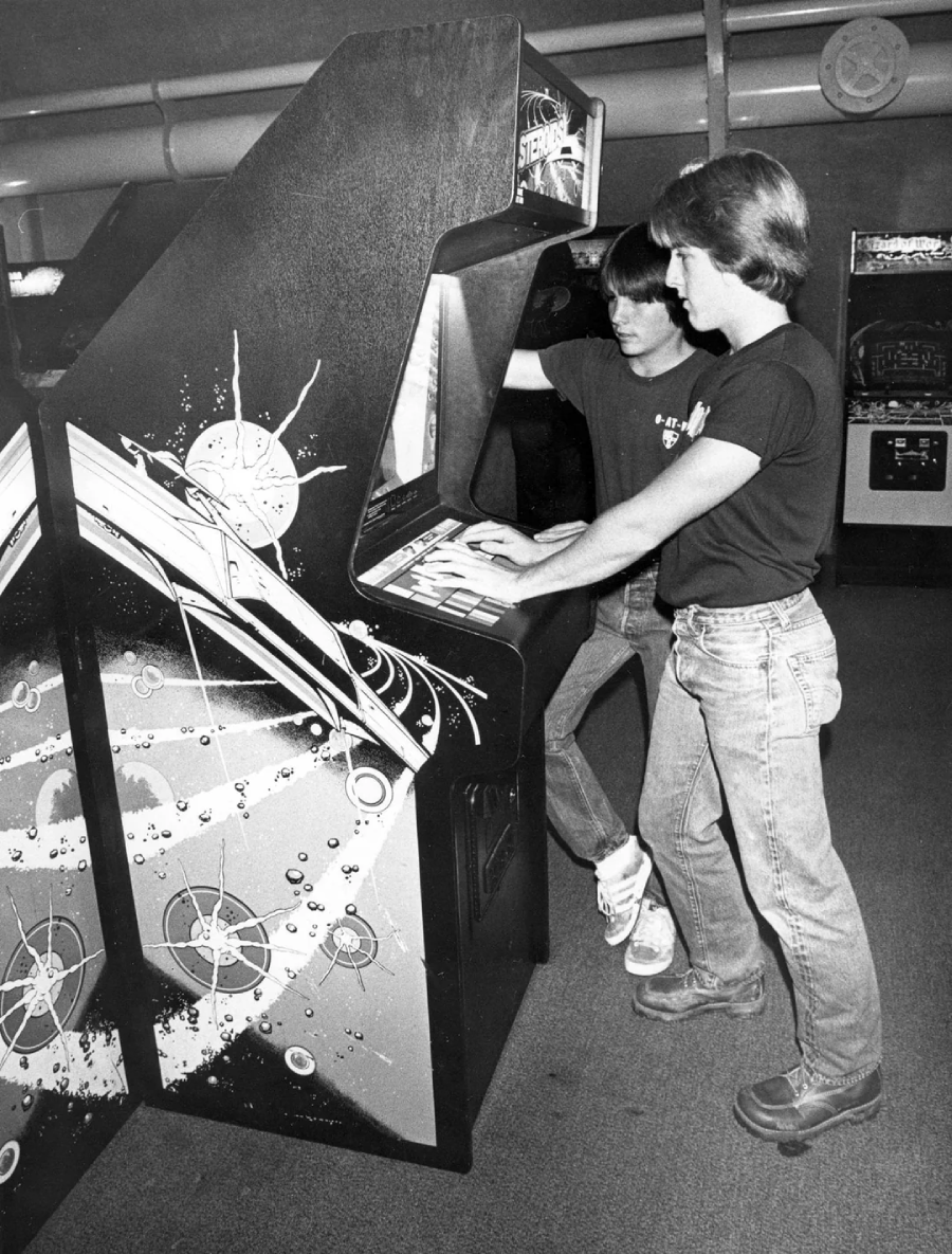Two young men stand at a vintage arcade game machine, focused on playing. The machine has a space-themed design with stars and planets. One player presses the buttons while the other watches. Retro arcade cabinets are visible in the background.