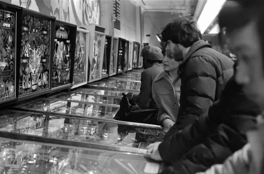 People stand side by side playing pinball machines in a brightly lit arcade. The scene is in black and white, capturing a vintage, nostalgic atmosphere.