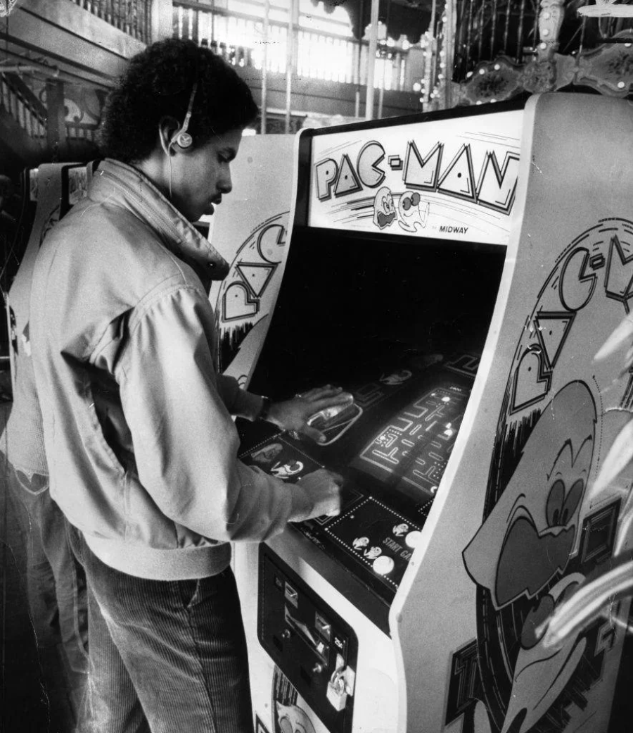 A person wearing headphones and a jacket plays a Pac-Man arcade machine in a retro arcade setting. The black-and-white image shows the Pac-Man game screen and detailed artwork on the arcade cabinet.