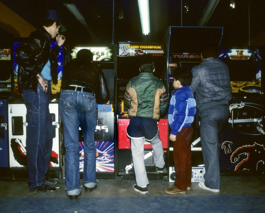 Five people, seen from behind, stand and play at vintage arcade game machines in a dimly lit arcade. They are dressed in 1980s-style jackets and jeans, with a child among the group.