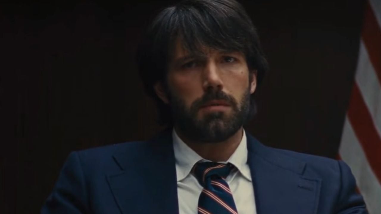 A man with dark hair and a beard, wearing a navy suit and striped tie, sits in front of a blurred American flag. He has a serious expression and is facing forward.