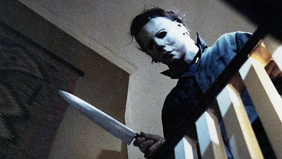 A person wearing a white mask and dark clothing stands at the top of a staircase, holding a large kitchen knife in a menacing manner, viewed from a low angle.