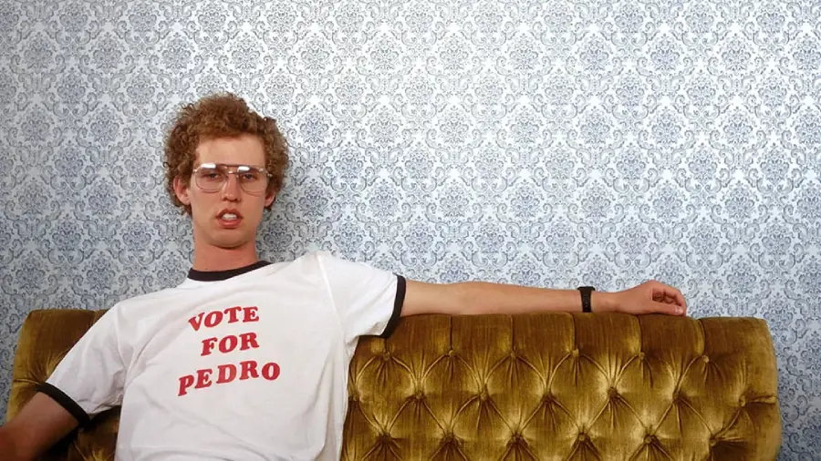A man with curly hair and glasses sits on a gold tufted sofa, wearing a white T-shirt that says "VOTE FOR PEDRO" in red letters, against a patterned wallpaper background.