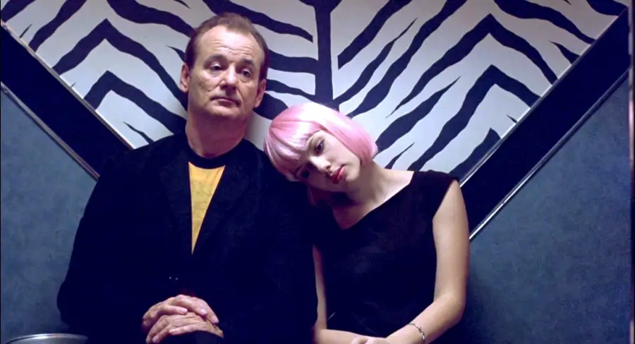 A man in a dark jacket sits next to a woman with a pink bob haircut resting her head on his shoulder, both against a wall with a black-and-white zebra print pattern.
