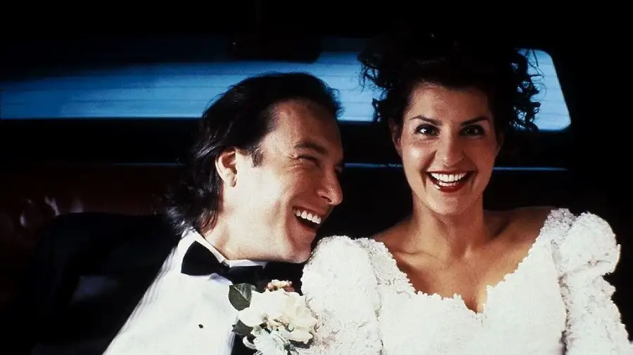 A bride in a white dress and a groom in a tuxedo sit close together, smiling and laughing happily, inside the back of a car.