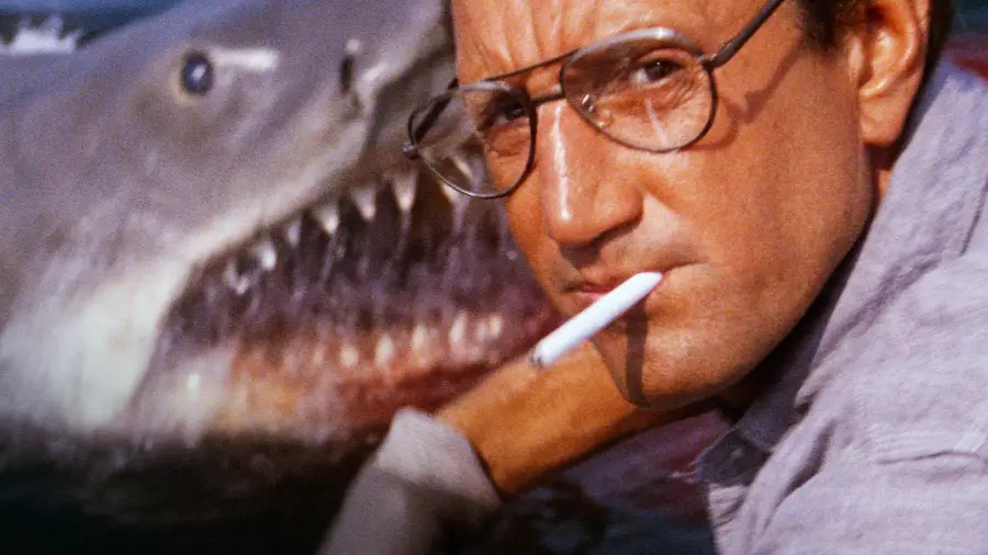 A man with glasses and a cigarette in his mouth looks intently ahead as a large shark with an open mouth approaches from behind in the water.