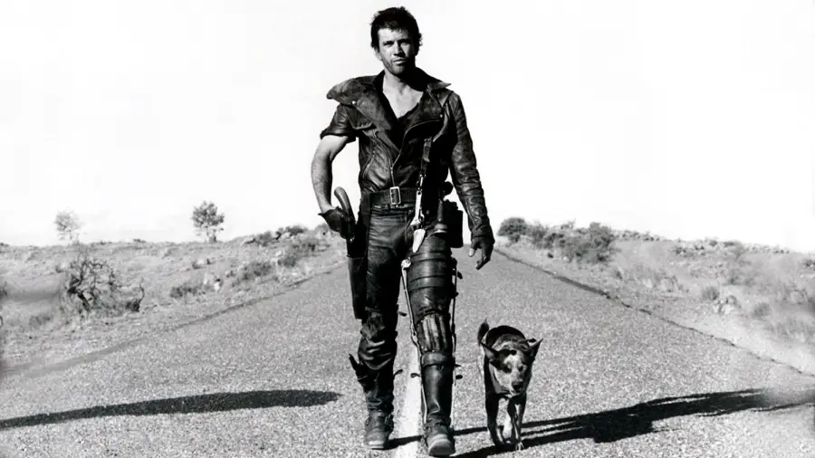 A man in rugged clothing and armor walks down an empty road in a barren landscape, holding a shotgun and accompanied by a dog. The image is in black and white, evoking a post-apocalyptic atmosphere.