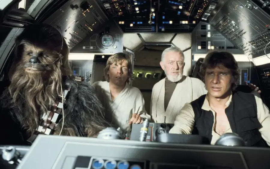 Four characters from Star Wars—Chewbacca, Luke Skywalker, Obi-Wan Kenobi, and Han Solo—sit in the cockpit of the Millennium Falcon, surrounded by control panels and lights.