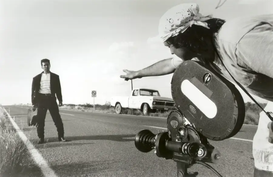 A black-and-white photo shows a man in a suit standing on a deserted road, while a filmmaker with a headscarf points toward him behind a vintage film camera. An old pickup truck is parked in the background.