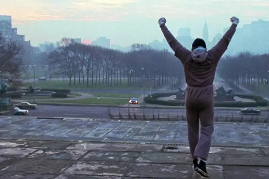 A person in a gray tracksuit stands triumphantly at the top of outdoor stone steps with arms raised, overlooking a city park at sunrise.