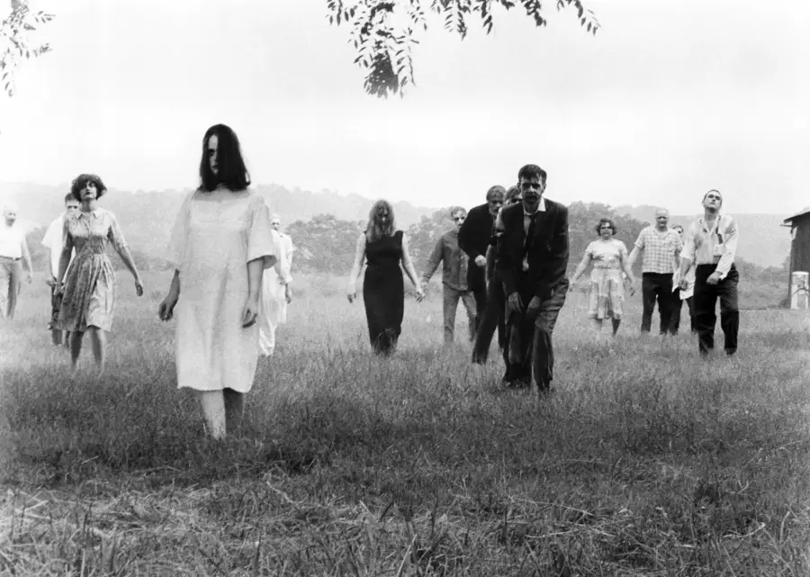 A group of people in tattered clothing walk across a grassy field with blank expressions, resembling zombies; the scene is in black and white, with trees and a building in the background.