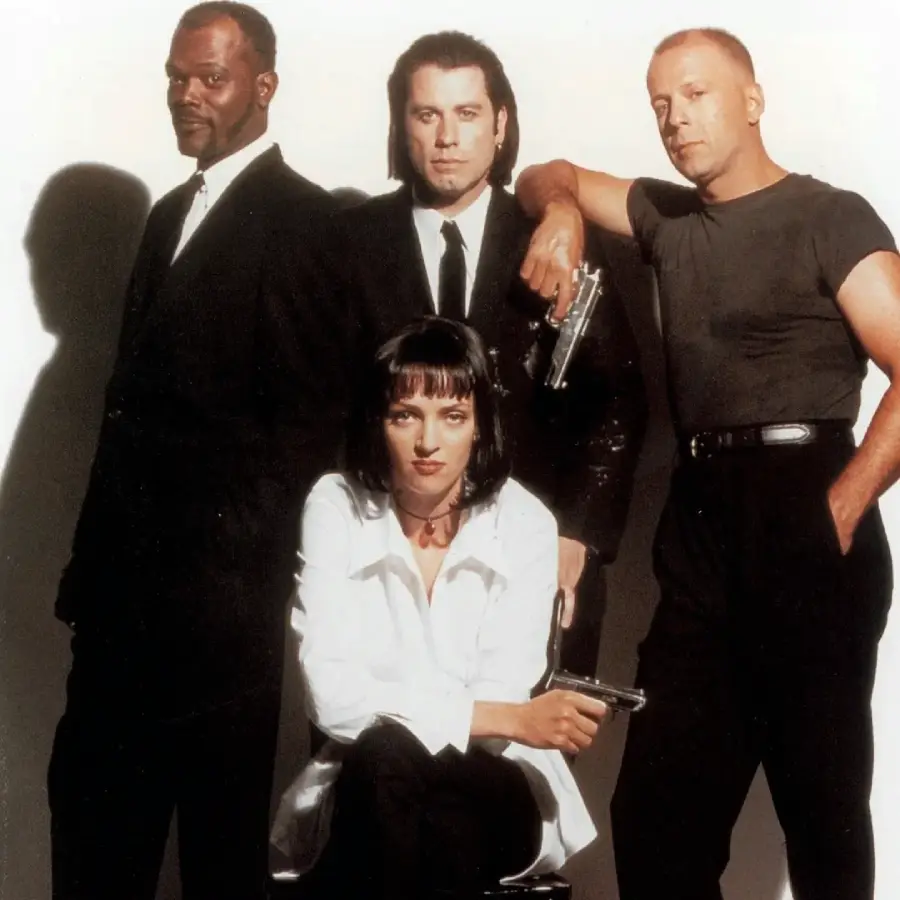 Four people pose against a white background; three men stand behind a woman sitting on a chair. The men wear dark clothing, while the woman wears a white shirt, all holding serious expressions.