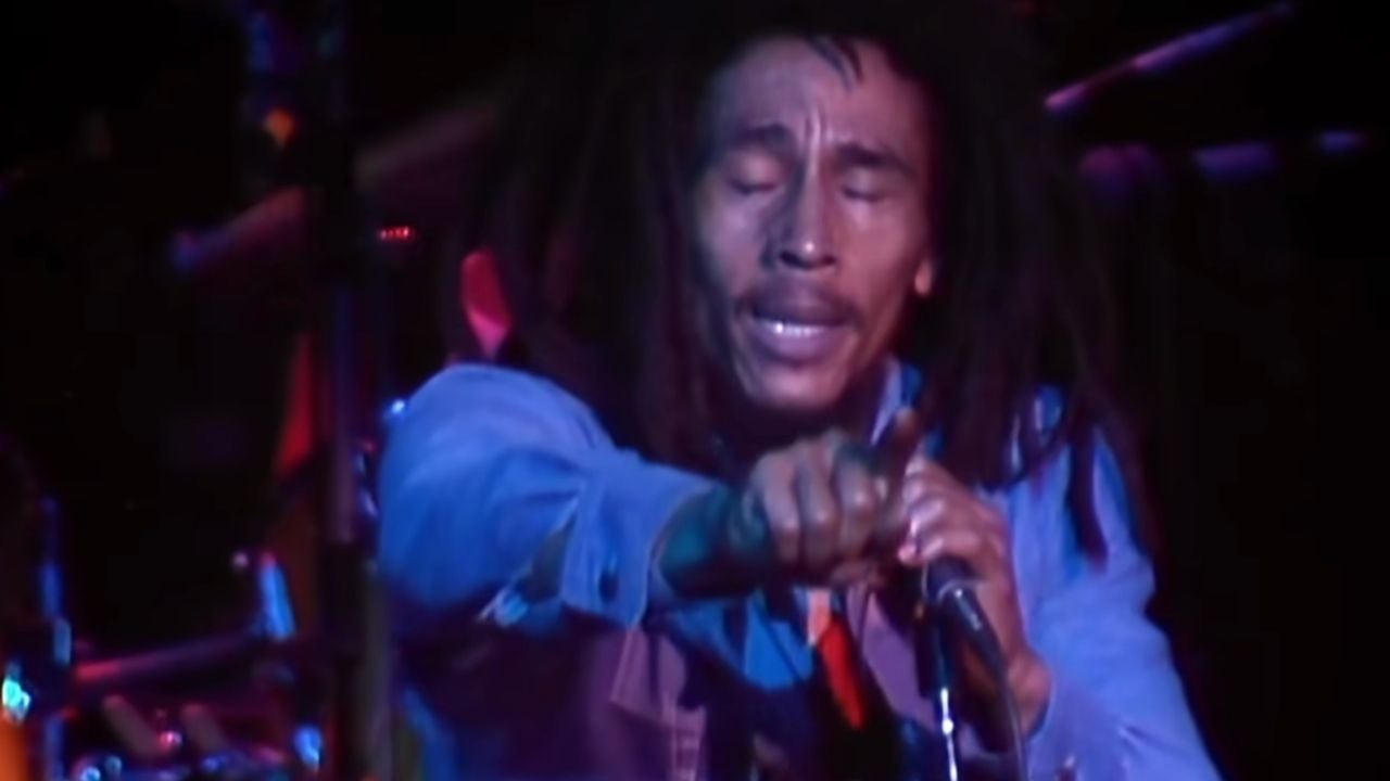 A man with dreadlocks sings passionately into a microphone on stage, wearing a denim jacket, with colored stage lights illuminating his face.
