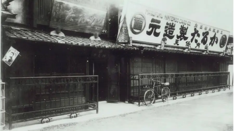 Black-and-white photo of an old Japanese building with signs in Japanese above; a bicycle is parked outside, and wooden railings front the structure. The scene appears historical.