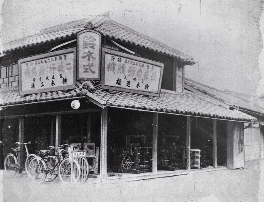 Black and white photo of a traditional Japanese building with a tiled roof, large signs in Japanese, glass windows, and several bicycles parked outside. Old machinery can be seen inside the shop.