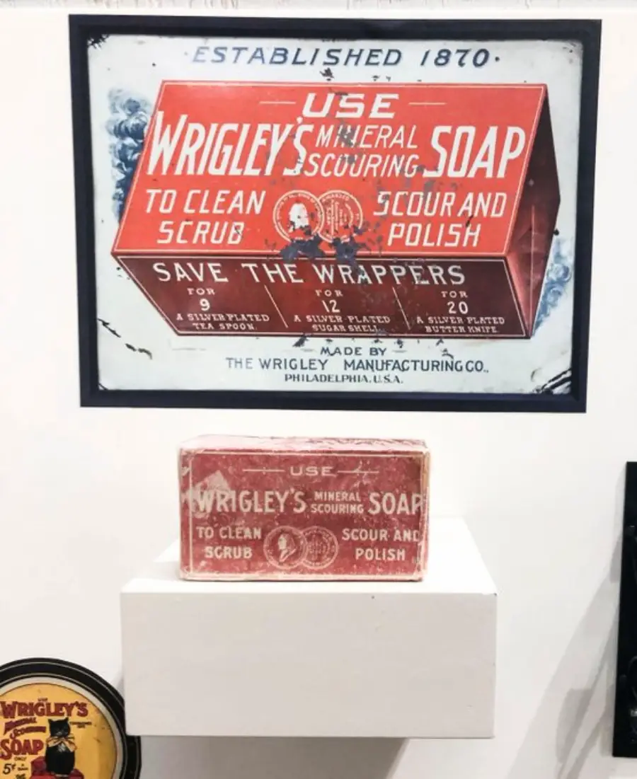 A vintage Wrigley’s Mineral Scouring Soap box is displayed on a white pedestal, with a matching old advertisement poster above it showing usage instructions and promotional text in bold red and white lettering.