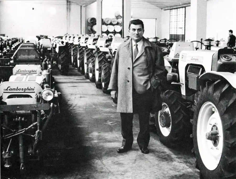 A man in a suit and overcoat stands in a warehouse next to rows of Lamborghini tractors, with more tractors lined up behind him. The setting appears industrial, with high ceilings and large windows.