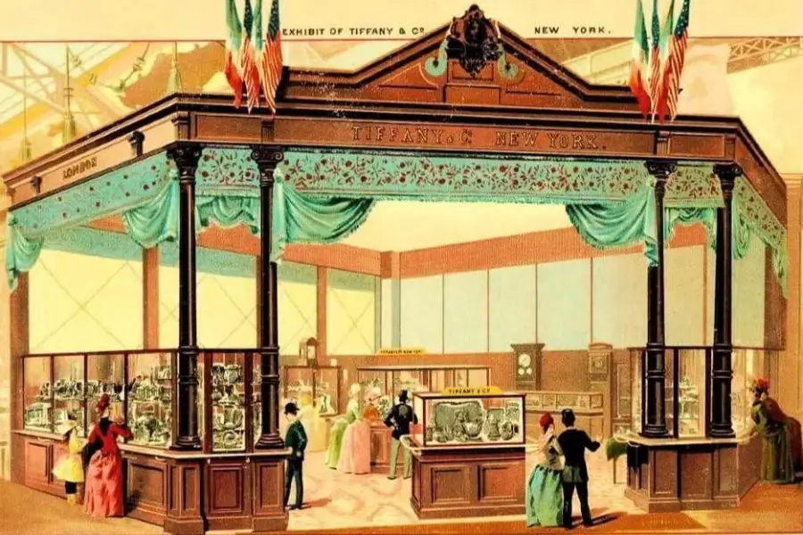 Illustration of a Tiffany & Co. exhibit at a fair, showing an ornate booth with flags, teal drapery, and glass display cases. Visitors in 19th-century attire view the showcased jewelry and items.