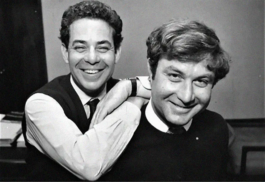 Two men smiling, one with curly dark hair and a vest resting his arm on the shoulder of the other man with lighter, wavy hair and a sweater. Both are looking happily at the camera in a black-and-white photo.
