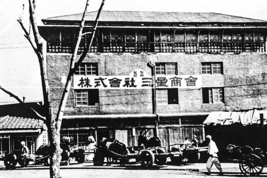 Black-and-white photo of an old three-story brick building with a Japanese sign, several rickshaws parked in front, and people walking or standing nearby on the street. A bare tree is visible in the foreground.