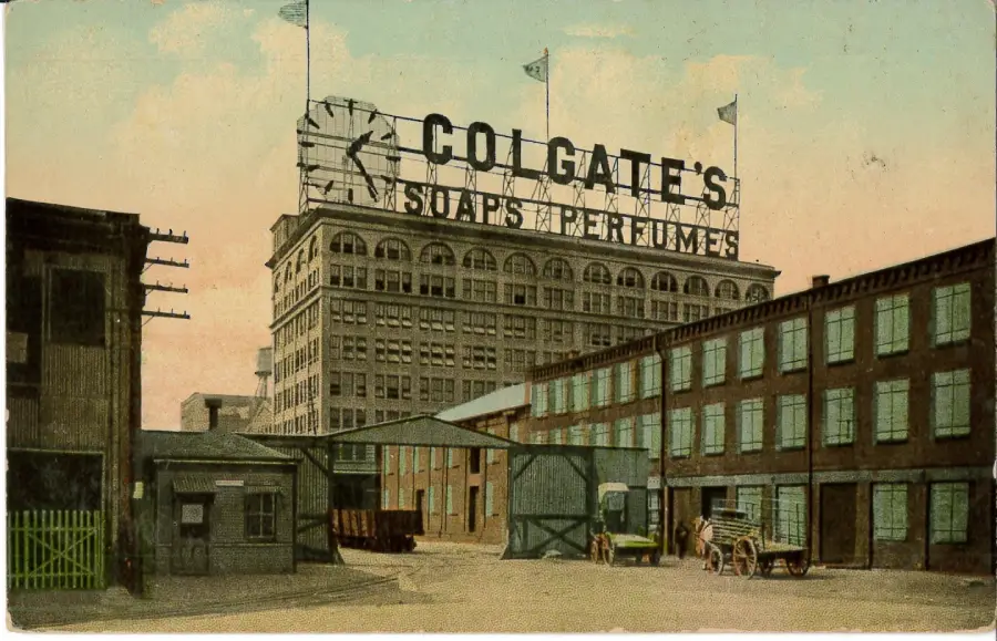 Historic postcard image of Colgate's factory, featuring a large building with a rooftop sign reading "Colgate's Soaps & Perfumes," a clock, and carts in the courtyard below.