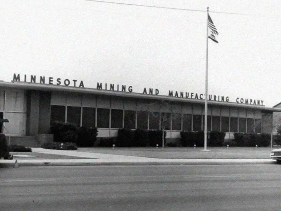 A black-and-white photo shows a low, rectangular building with "Minnesota Mining and Manufacturing Company" on its roof. An American flag flies on a tall pole near the entrance. Bushes line the front.