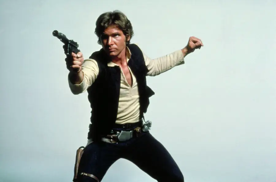 A man in a black vest, beige shirt, and dark pants strikes an action pose, aiming a blaster pistol with one arm extended and the other bent. He has brown hair and wears a holster on his hip, standing against a plain background.