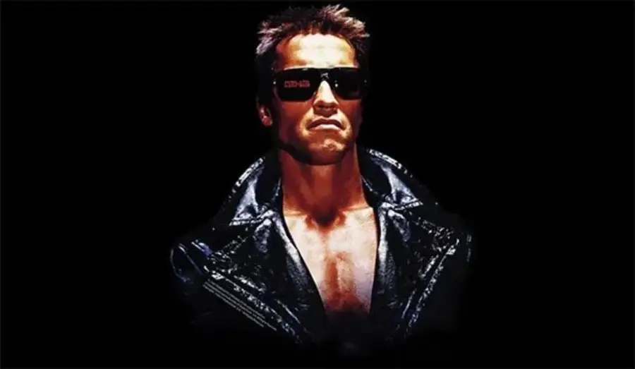 A man wearing dark sunglasses and a black leather jacket stands against a black background, with a tough expression and chest partially exposed.