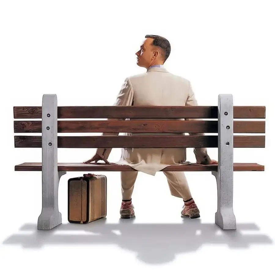 A man in a light-colored suit sits on a wooden bench, facing away, with a suitcase placed on the ground beside him against a plain white background.