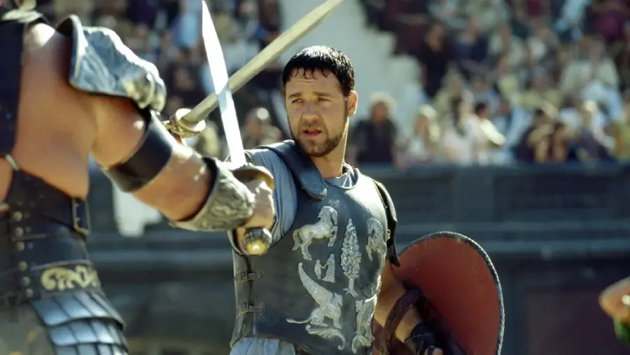 A man in Roman armor holds a sword and round shield, facing another armored opponent in an arena, with a blurred crowd watching in the background.