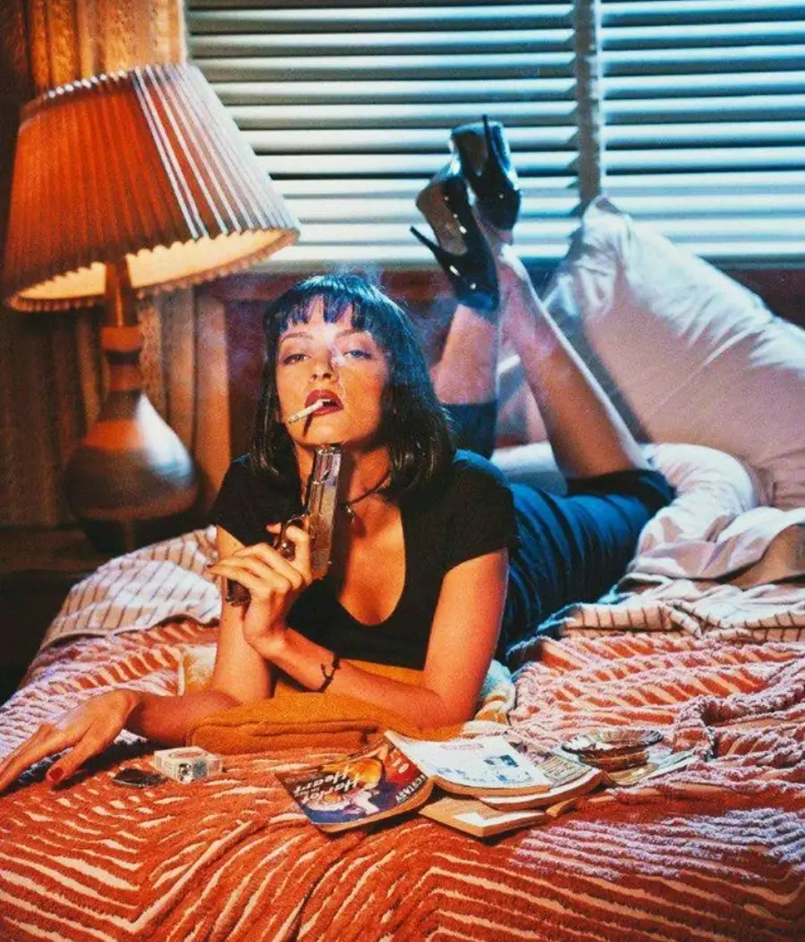 A woman with short dark hair in a black dress lies on a bed, holding a gun to her lips. She is surrounded by books, magazines, and cigarettes, with a vintage lamp and blinds in the background.