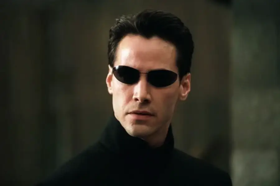 A person with short dark hair wearing black sunglasses and a high-collared black coat stands against a blurred background, looking serious and focused.