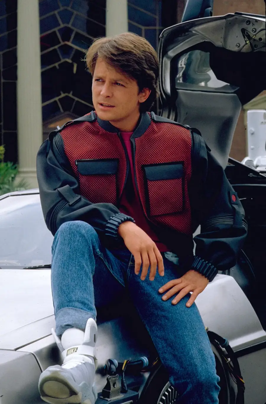 A young man in a red and black jacket and blue jeans sits on the edge of a futuristic silver car with gull-wing doors, looking off to the side with one knee raised and his hand resting on his leg.